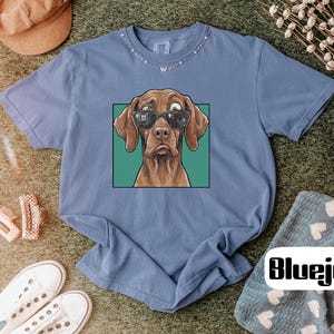 May include: A blue short-sleeve t-shirt featuring a graphic print of a brown dog wearing black sunglasses. The dog is centred within a green square frame. The shirt has a crew neckline and a relaxed fit, showcasing a casual and simple design.
