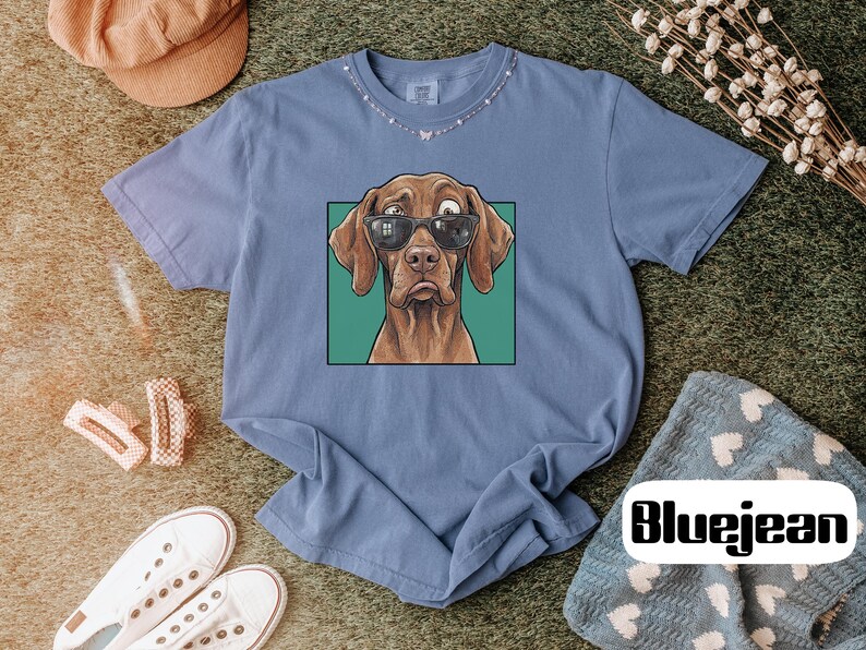 May include: A blue short-sleeve t-shirt featuring a graphic print of a brown dog wearing black sunglasses. The dog is centred within a green square frame. The shirt has a crew neckline and a relaxed fit, showcasing a casual and simple design.