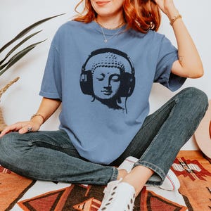 May include: A dusty blue short-sleeve t-shirt featuring a black graphic print of a Buddha head wearing large over-ear headphones. The shirt has a crew neckline and a relaxed fit, paired with dark blue denim jeans and white sneakers.