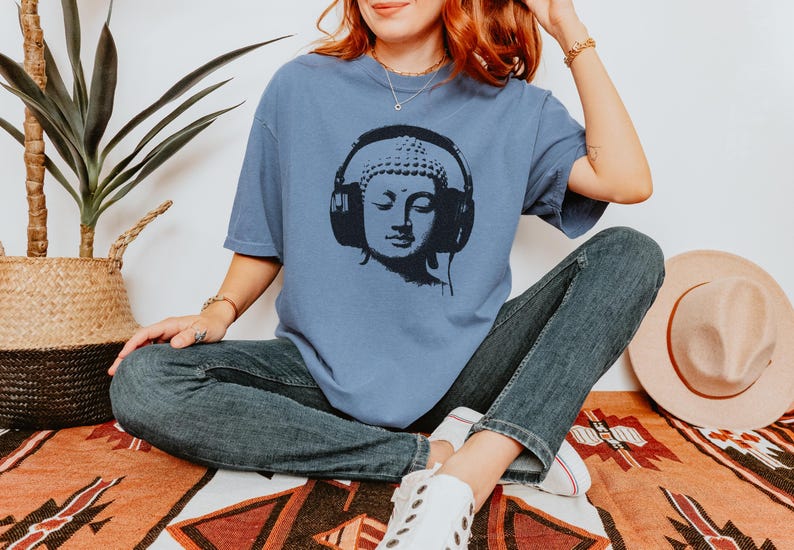 May include: A dusty blue short-sleeve t-shirt featuring a black graphic print of a Buddha head wearing large over-ear headphones. The shirt has a crew neckline and a relaxed fit, paired with dark blue denim jeans and white sneakers.
