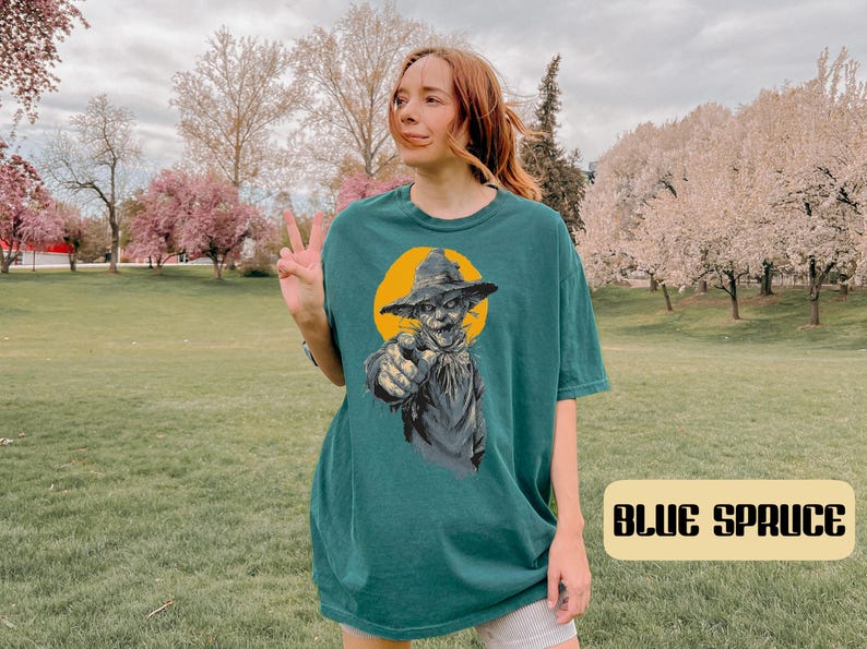 May include: A dark teal short-sleeve t-shirt featuring a graphic print of a scarecrow wearing a hat and pointing forward against a yellow circular background. The text "BLUE SPRUCE" is printed in a bold, dark font on a light rectangular label.