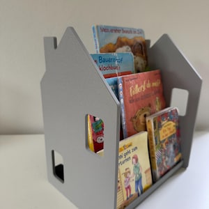 Könnte beinhalten: Ein hellgraues, hausförmiges Bücherregal mit ausgeschnittenen Fenstern, das mehrere Kinderbücher mit bunten Einbänden enthält, darunter die Titel Mein erster Besuch im Zoo, Bauernhof, Fütterst du mich?, Unkaputtbar und Ein Tag in der Stadt.