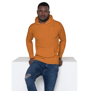 Peut inclure: Un sweat-shirt à capuche orange brûlé uni avec une poche kangourou sur le devant, des manches longues à poignets côtelés et une petite icône blanche brodée sur la poitrine gauche, associé à un jean bleu présentant une déchirure au genou droit.