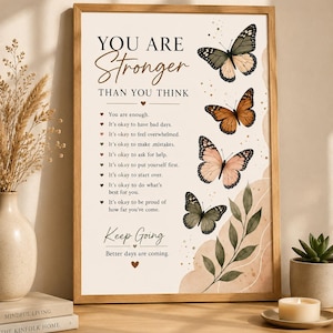 Könnte beinhalten: Ein gerahmter Kunstdruck mit dem Text "YOU ARE Stronger THAN YOU THINK", einer Liste von Affirmationen und dem Satz "Keep Going. Better days are coming." Das Design zeigt vier Schmetterlinge und eine Illustration eines Blattzweigs.