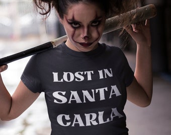 May include: A woman wearing a dark gray t-shirt with the text "LOST IN SANTA CARLA" in white letters. She is holding a wooden baseball bat over her shoulder and has clown makeup on.