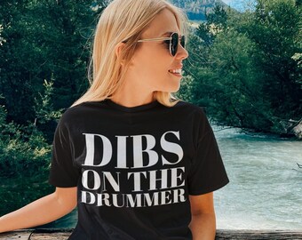 Puede incluir: Una camiseta negra con texto blanco que dice "DIBS ON THE DRUMMER". La persona que lleva la camiseta está de pie en un puente de madera sobre un río.