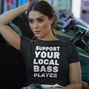 May include: A woman wearing a black t-shirt with the text "Support Your Local Bass Player" in white lettering. She is sitting in a black and green amusement park ride.