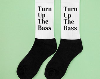 以下が含まれることがあります： 「Turn Up The Bass」の文字が黒い太字でプリントされた、白と黒の靴下のペア。靴下は白い上部と黒い足の部分があります。背景は明るい緑色です。