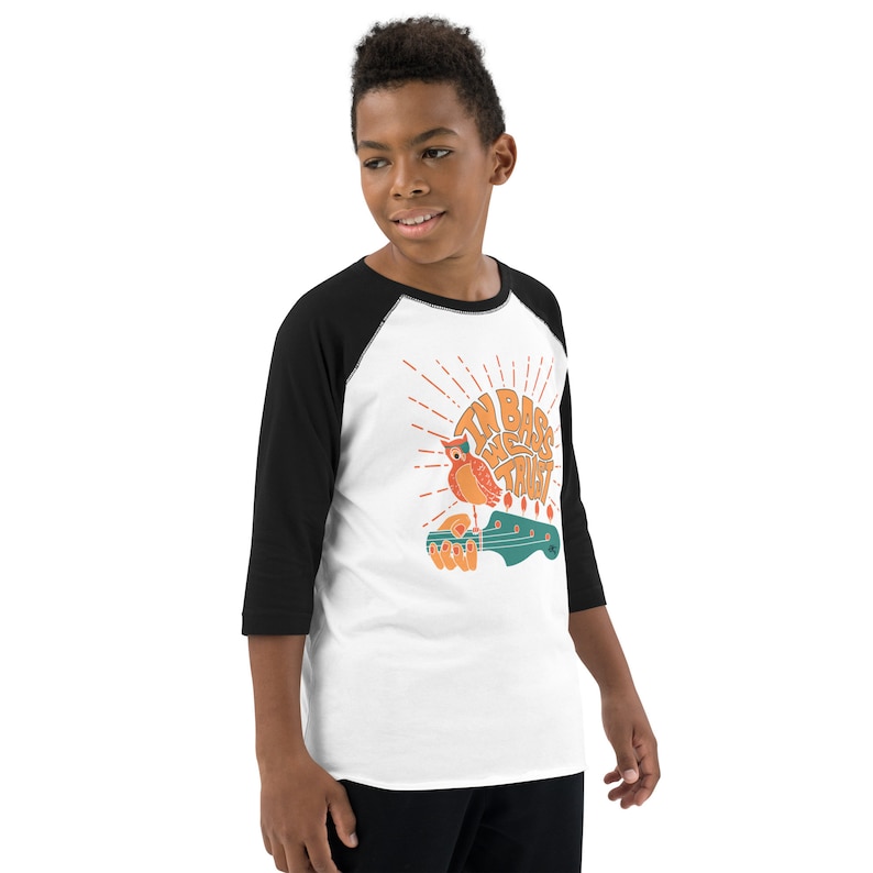 May include: A white and black baseball tee shirt with a graphic of an owl perched on a bass guitar. The text on the shirt reads "In bass we trust".