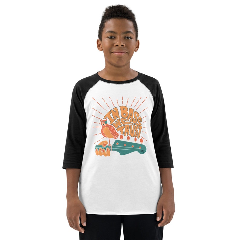 May include: A white and black baseball tee shirt with a graphic of a bird perched on a bass guitar. The graphic says "In Bass We Trust" with a sunburst behind the image.