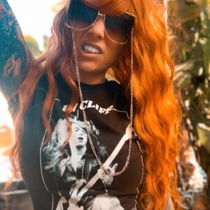 Puede incluir: Una mujer con el pelo largo y de color naranja brillante lleva una camiseta negra con un gr&aacute;fico blanco de una persona tocando la guitarra. La camiseta tiene el texto "We Trust" y "Cliff". Lleva gafas de sol con una cadena dorada.