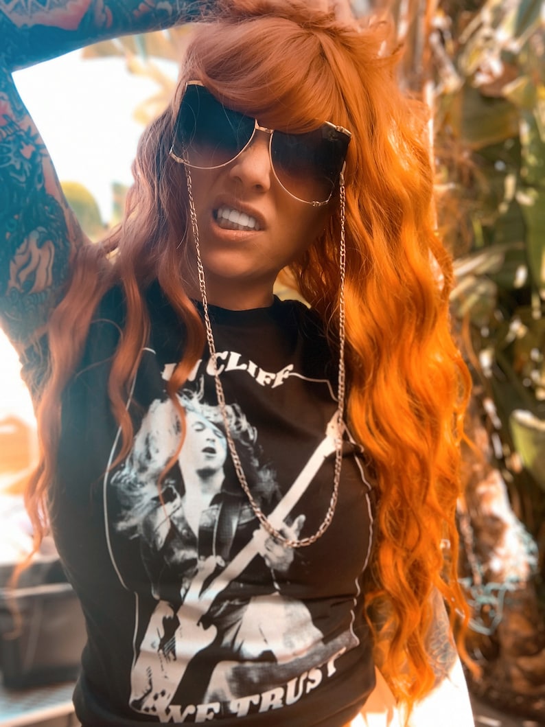 Puede incluir: Una mujer con el pelo largo y de color naranja brillante lleva una camiseta negra con un gr&aacute;fico blanco de una persona tocando la guitarra. La camiseta tiene el texto "We Trust" y "Cliff". Lleva gafas de sol con una cadena dorada.