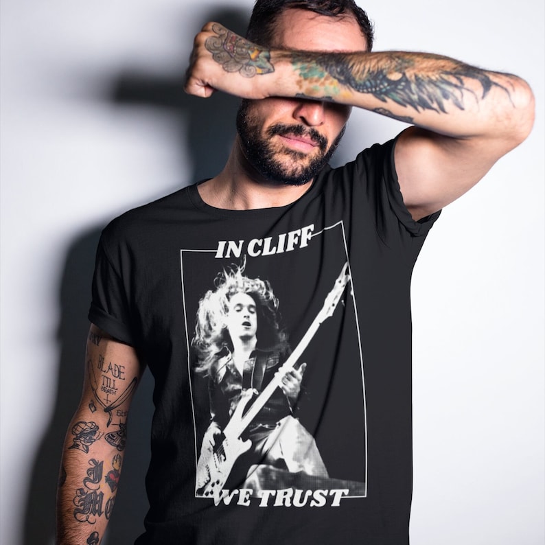Puede incluir: Una camiseta negra con un gr&aacute;fico blanco de un m&uacute;sico tocando una guitarra. El gr&aacute;fico incluye el texto "IN CLIFF WE TRUST".