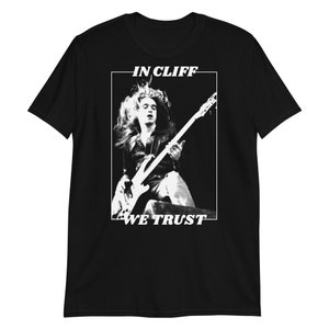 Puede incluir: Imagen en blanco y negro de un m&uacute;sico tocando un bajo. El texto "IN CLIFF WE TRUST" est&aacute; impreso en la camiseta.