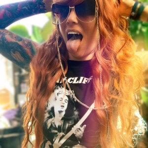 Puede incluir: Una mujer con el pelo largo y ondulado de color rojo lleva una camiseta negra con un gr&aacute;fico blanco de una persona tocando la guitarra. La camiseta tiene el texto "INCLIF" y "WE TRUST". Lleva gafas de sol con una cadena dorada.