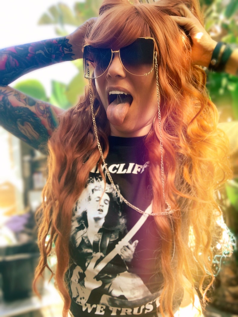 Puede incluir: Una mujer con el pelo largo y ondulado de color rojo lleva una camiseta negra con un gr&aacute;fico blanco de una persona tocando la guitarra. La camiseta tiene el texto "INCLIF" y "WE TRUST". Lleva gafas de sol con una cadena dorada.
