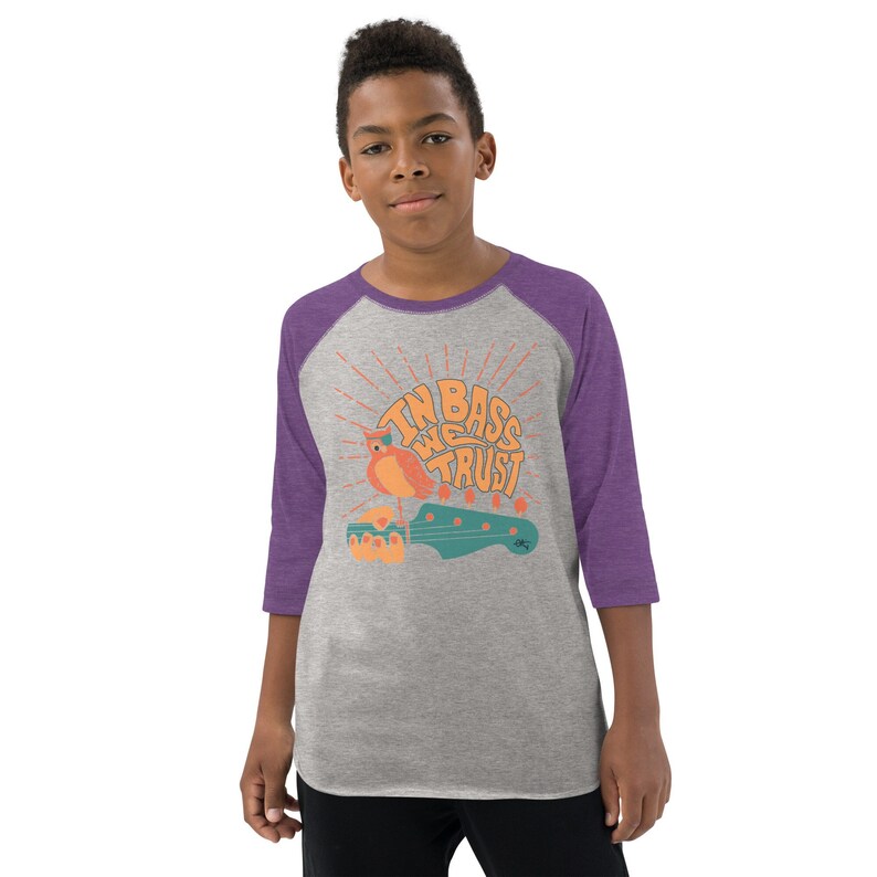 May include: A grey and purple youth baseball t-shirt with a graphic of a bass guitar and a bird with the text "In Bass We Trust".
