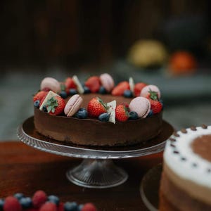 Puede incluir: Un pastel de chocolate redondo cubierto con fresas rojas frescas, arándanos y macarons de color rosa claro. El pastel está decorado con pequeños trozos de chocolate blanco y se asienta sobre un soporte de cristal. Un segundo pastel es visible al frente.
