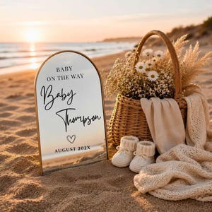 以下が含まれることがあります： BABY ON THE WAY、Baby Thompson、AUGUST 202Xとハートのアイコンが黒い文字で書かれたアーチ型のミラーサイン。その横には、白い花とパンパスグラスが入った編み込みの籐かごがあり、クリーム色のニットブランケットとベビーシューズが掛けられています。