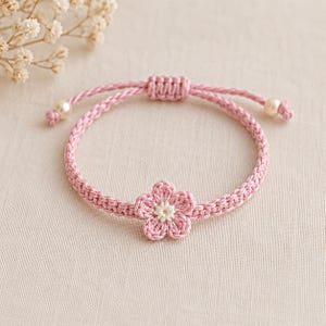 May include: A pink braided cord bracelet featuring a central five-petaled flower charm with a white center. The bracelet includes an adjustable sliding knot closure and two small cream-colored beads at the ends of the cord strings.