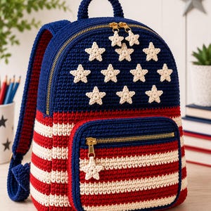 May include: A crochet backpack featuring a dark blue upper section adorned with eleven cream-colored stars. The lower portion and front pocket display horizontal red and cream stripes. It includes gold-toned zippers with star-shaped zipper pulls.