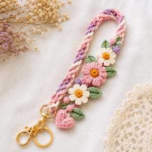May include: A braided crochet wristlet strap in shades of pink, purple, and white, adorned with crocheted flowers, green leaves, and a small pink heart. The strap features a gold-toned metal lobster clasp and a circular ring for attachment.