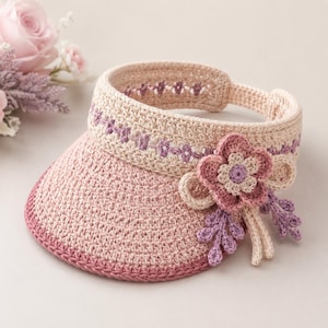 May include: A crochet sun visor in shades of beige and dusty pink, featuring a wide brim with a darker pink edge. The headband is decorated with a purple stitched pattern and a large, layered crochet flower accent in pink, beige, and purple.