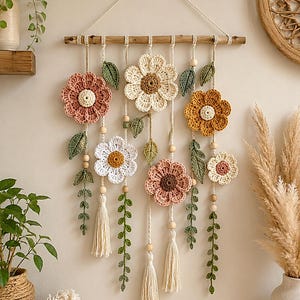 May include: A wall hanging featuring crocheted flowers in shades of pink, white, and mustard yellow, accented with green crocheted leaves and vines. The piece is suspended from a wooden branch by cream-colored cords with wooden beads and tassels.