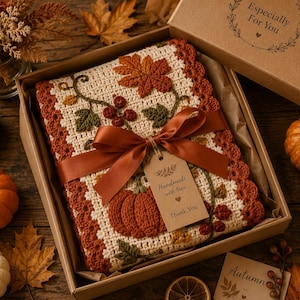 May include: A rectangular crochet blanket featuring an off-white base with a rust-colored border, decorated with crocheted autumn leaves, berries, and a pumpkin. The blanket is tied with a brown satin ribbon and a tag labeled Handmade with love.