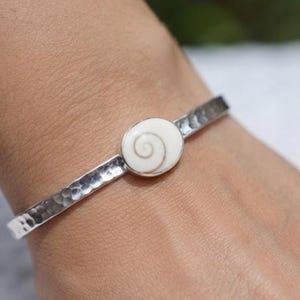 Può includere: Un bracciale a fascia color argento caratterizzato da una banda metallica martellata e testurizzata, con un inserto centrale in conchiglia bianca di forma ovale decorato da un motivo a spirale naturale. Il bracciale presenta un design aperto e una finitura lucida, evidenziando un netto contrasto tra la superficie metallica e l'elemento organico.