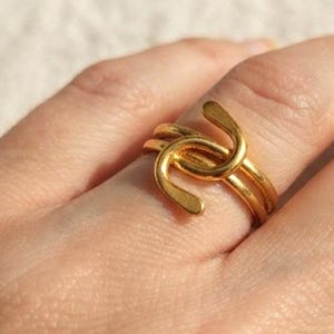 Può includere: Un anello color oro caratterizzato da un design in filo metallico continuo e sinuoso che si ripiega su se stesso. La fascia è realizzata in un unico pezzo di metallo lucido color oro, creando una forma aperta e sovrapposta sul dito.