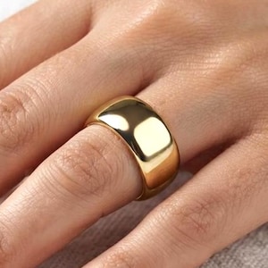 May include: A wide, polished gold band ring with a smooth, reflective surface and a rounded profile. The ring features a solid, continuous construction with a high-shine finish that catches the light, creating bright highlights across the metal.