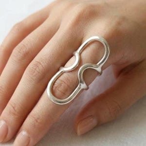May include: A silver-toned ring featuring an elongated, double-loop design that spans across two finger joints. The metal band is smooth and continuous, forming two connected oval shapes that sit vertically along the length of the finger.