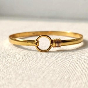Op de afbeelding: Een goudkleurige armband met een centrale cirkelvormige ring als sluiting. De armband heeft een gladde, gepolijste afwerking en bevat een klein detail van gewikkeld draad naast het cirkelvormige sluitpunt.