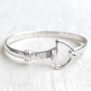 May include: A polished silver-tone bangle bracelet featuring a central equestrian-inspired bit design. The metal band is smooth and rounded, transitioning into a triangular bit element with a small hook closure detail on one side of the frame.