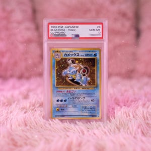 May include: A 1999 Japanese Blastoise Holo CD Promo trading card encased in a clear protective slab. The card features the blue water-type creature with cannons on its back, Japanese text, and a Gem Mint 10 grade label at the top of the case.