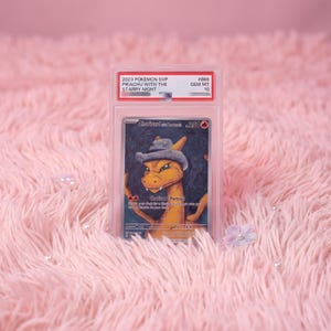 May include: A graded 2023 Pokemon SVP Charizard trading card in a clear plastic slab. The card features an orange dragon-like creature wearing a grey hat against a dark blue swirling background. The label reads 2023 POKEMON SVP PIKACHU WITH THE STARRY NIGHT #666 GEM MT 10.