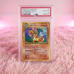 Könnte beinhalten: Eine japanische Charizard-Holo CD Promo Sammelkarte aus dem Jahr 1999, bewertet mit GEM MT 10, in einer durchsichtigen Kunststoffhülle. Die Karte zeigt eine holografische Illustration des orangefarbenen Drachen mit blauen Flügeln.