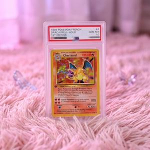 Puede incluir: Una carta coleccionable holográfica Charizard de 1999, 1ª edición francesa, en una funda protectora transparente PSA. La etiqueta dice 1999 POKEMON FRENCH DRACAUFEU - HOLO 1ST EDITION #4 GEM MT 10. La carta muestra una ilustración de un dragón.