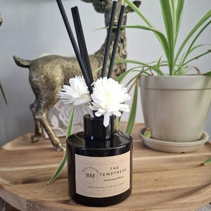Peut inclure: Un flacon diffuseur de parfum en verre noir avec une étiquette beige portant l'inscription Black Mustard BM The Temptress Home Reed Diffuser Hand Created in the New Forest. Il contient des bâtonnets noirs et deux fleurs décoratives blanches.