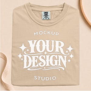 Può includere: Una t-shirt beige con la scritta bianca MOCKUP YOUR DESIGN STUDIO e stelle bianche. L'etichetta Comfort Colors all'interno del colletto indica una taglia large, realizzata in 100% cotone in Honduras.