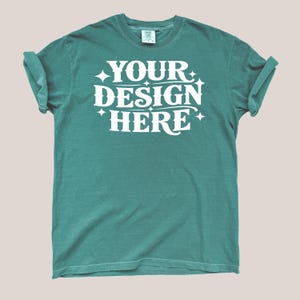 Könnte beinhalten: Ein kurzärmeliges T-Shirt in Blaugrün mit hochgekrempelten Ärmeln und Rundhalsausschnitt. In der Mitte des Shirts befindet sich der weiße Schriftzug YOUR DESIGN HERE, umgeben von kleinen vierzackigen Sternsymbolen. Markenetikett sichtbar.