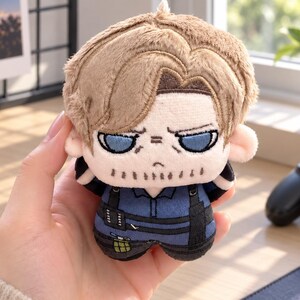 May include: A small plush toy featuring a character with light brown hair, blue eyes, and a stubble beard. The character wears a dark blue tactical shirt with black straps and a small green grenade accessory attached to the waist area.