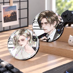 May include: Two circular pin-back buttons featuring anime-style illustrations of a person with blonde hair and a person with brown hair. Both buttons have a white background with black graphic accents, film strip motifs, and holographic star effects.