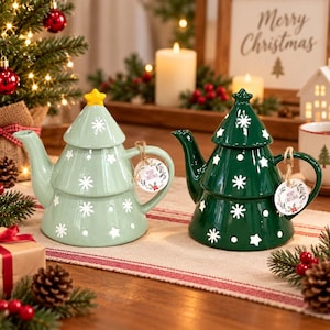 Puede incluir: Dos teteras de cerámica con forma de árbol de Navidad, una en verde claro y otra en verde oscuro. Ambas presentan patrones de copos de nieve y estrellas blancas, una estrella en la tapa y una etiqueta redonda atada al asa que dice Merry Christmas.