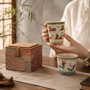Puede incluir: Una caja cuadrada con tapa color canela y exterior de tela con motivos circulares naranjas y marrones, acompañada de dos tazas de cerámica. Una taza muestra caballos marrones, mientras que la otra presenta figuras con túnicas azules y naranjas.