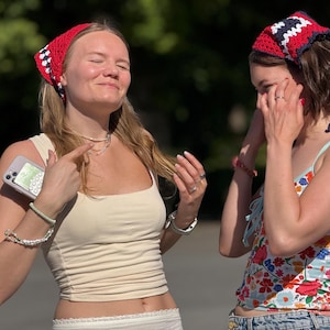 May include: Two crochet headscarves featuring a red, white, and navy blue geometric pattern. The headscarves are triangular in shape and tied at the back of the head, showcasing a textured yarn construction with a distinct colour-blocked design.