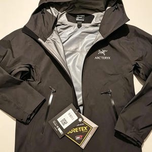 May include: A black Arc'teryx hooded jacket featuring a full-length front zipper, two zippered side pockets, and a light gray interior lining. The jacket includes a Gore-Tex brand tag attached to the front zipper pull and a white logo on the chest.