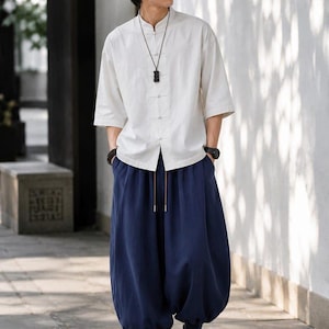 May include: A white short-sleeved shirt with a mandarin collar and front toggle closures paired with loose-fitting navy blue trousers featuring a drawstring waist and cuffed ankles. A black pendant necklace and a black wristwatch are also worn.