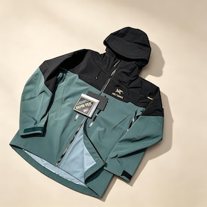 Op de afbeelding: Een tweekleurige jas met capuchon, voorzien van een zwart bovengedeelte en een gedempt groen ondergedeelte. De jas heeft een volledige ritssluiting aan de voorzijde, verstelbare manchetten en een Gore-Tex label op de borst. Het Arc'teryx-logo is goud geborduurd.